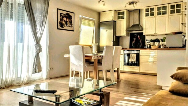 Ein modernes Wohnzimmer mit einem Glastisch, ein Essbereich mit weißen Stühlen und eine Küche mit weißen Schränken und Geräten.