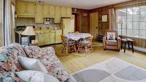 Una sala de estar acogedora con un sofá floral, una mesa de comedor con sillas, un sillón de cuero y un ventanal con vistas a árboles.