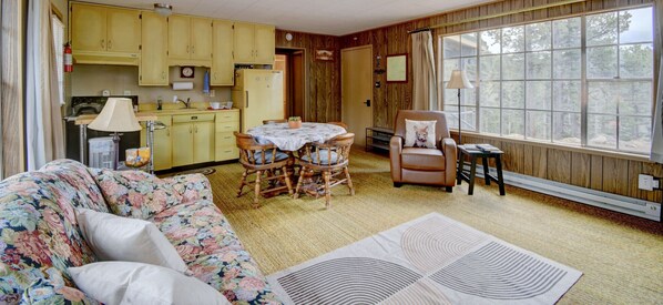 A cozy living room with a floral sofa, a dining table with chairs, a leather armchair, and a large window with a view of trees.