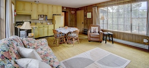 A cozy living room with a floral sofa, a dining table with chairs, a leather armchair, and a large window with a view of trees.