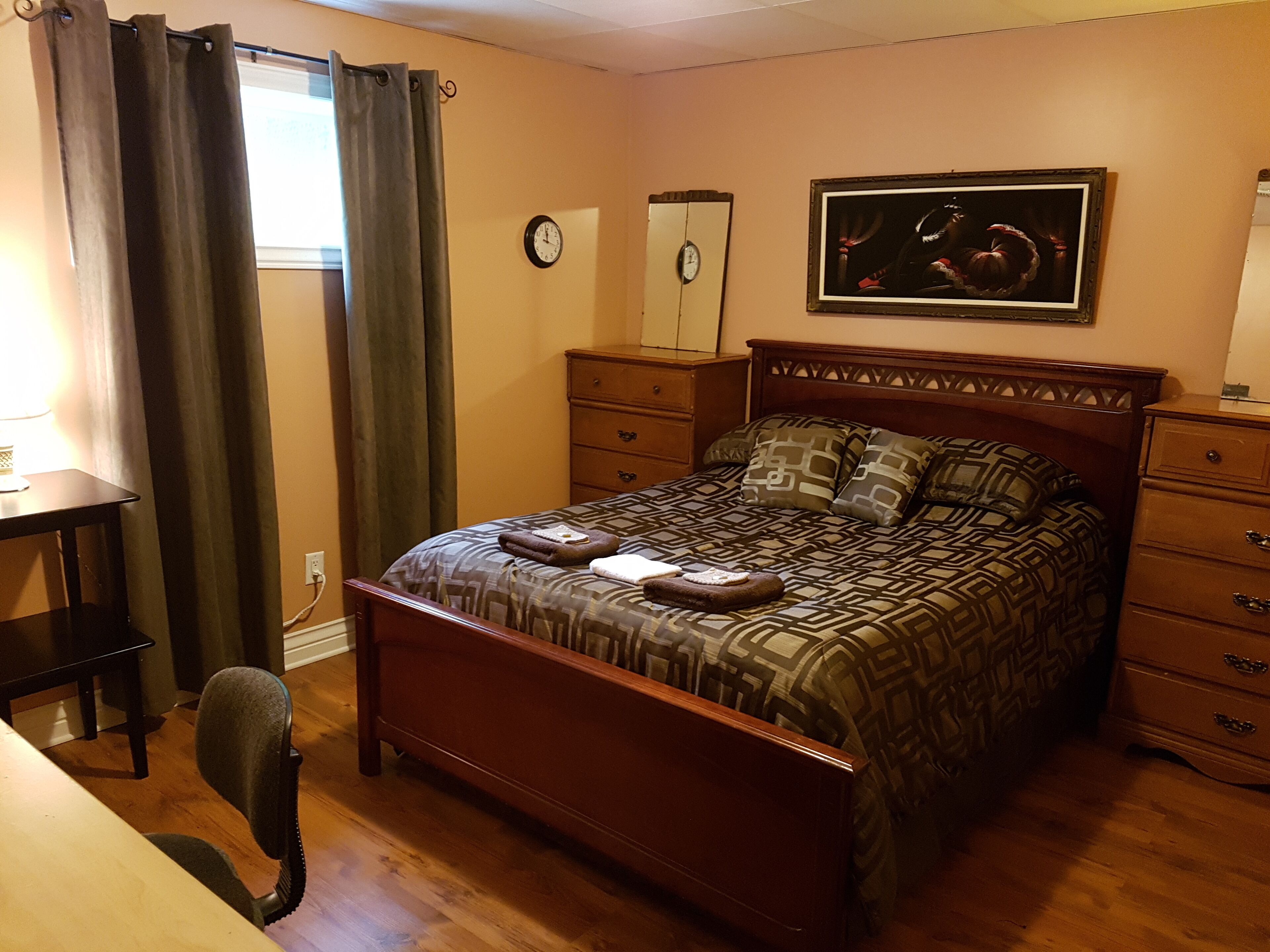 A bedroom with a wooden bed, a desk, a chair, a dresser, and a painting on the wall.