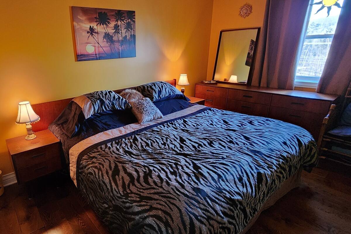 A bedroom with a zebra-patterned bedspread, a nightstand with a lamp, a wall-mounted fan, and a painting of a sunset.