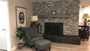 Un salon avec une cheminée en pierre, un fauteuil gris et une table d’appoint sur laquelle est posée une plante.