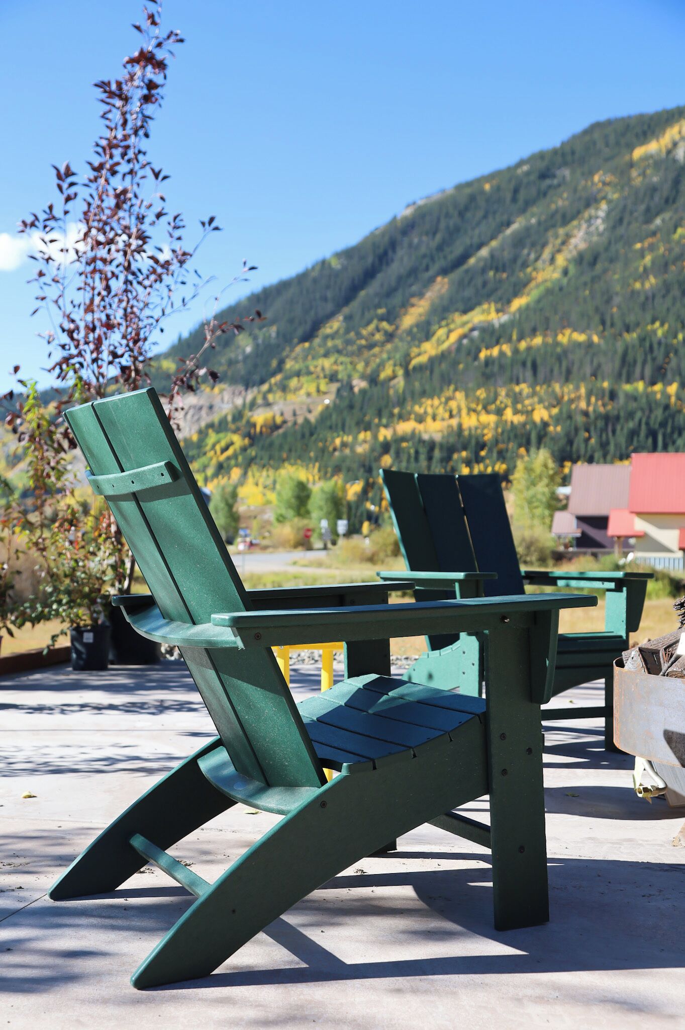 A green Adirondack chair with a metal frame, positioned on a concrete surface with a view of a mountain and a red-roofed building in the background.