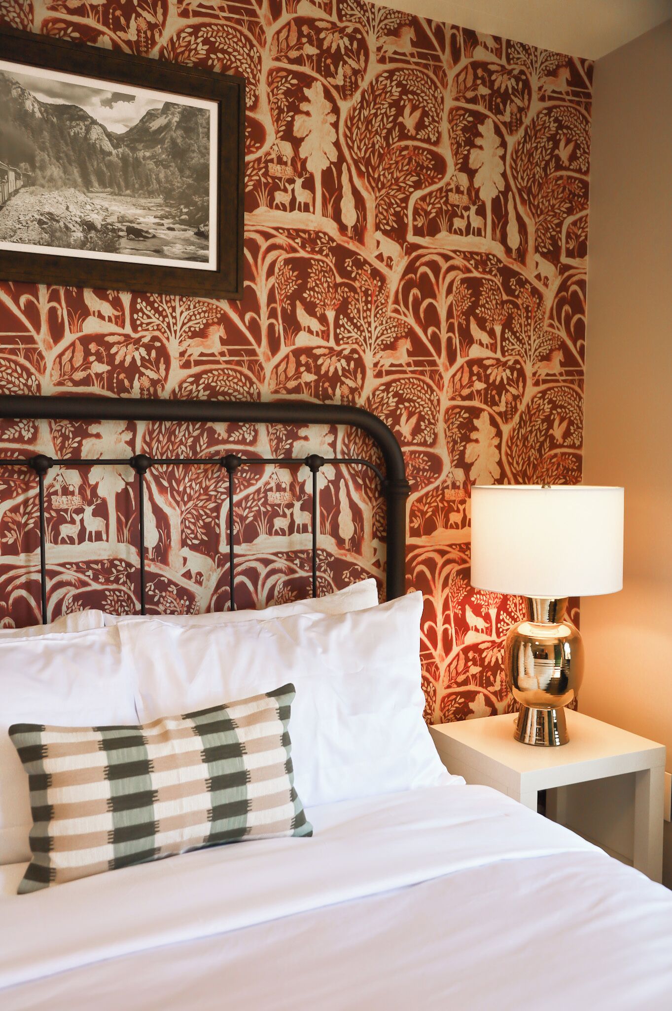 A bed with white linens, a checkered pillow, a metal headboard, a nightstand with a lamp, and a patterned wallpaper with a nature scene.