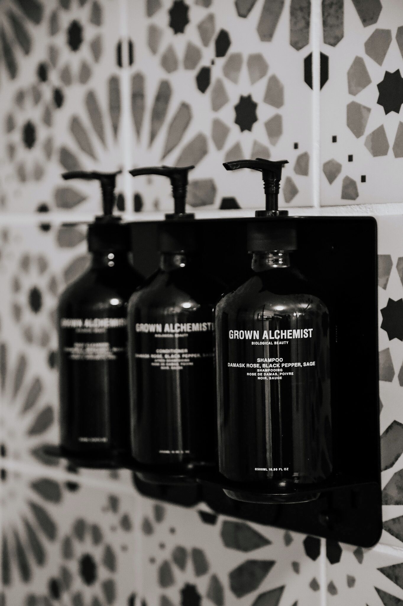 Three bottles of 'Grown Alchemist' shampoo on a shelf against a patterned wall.