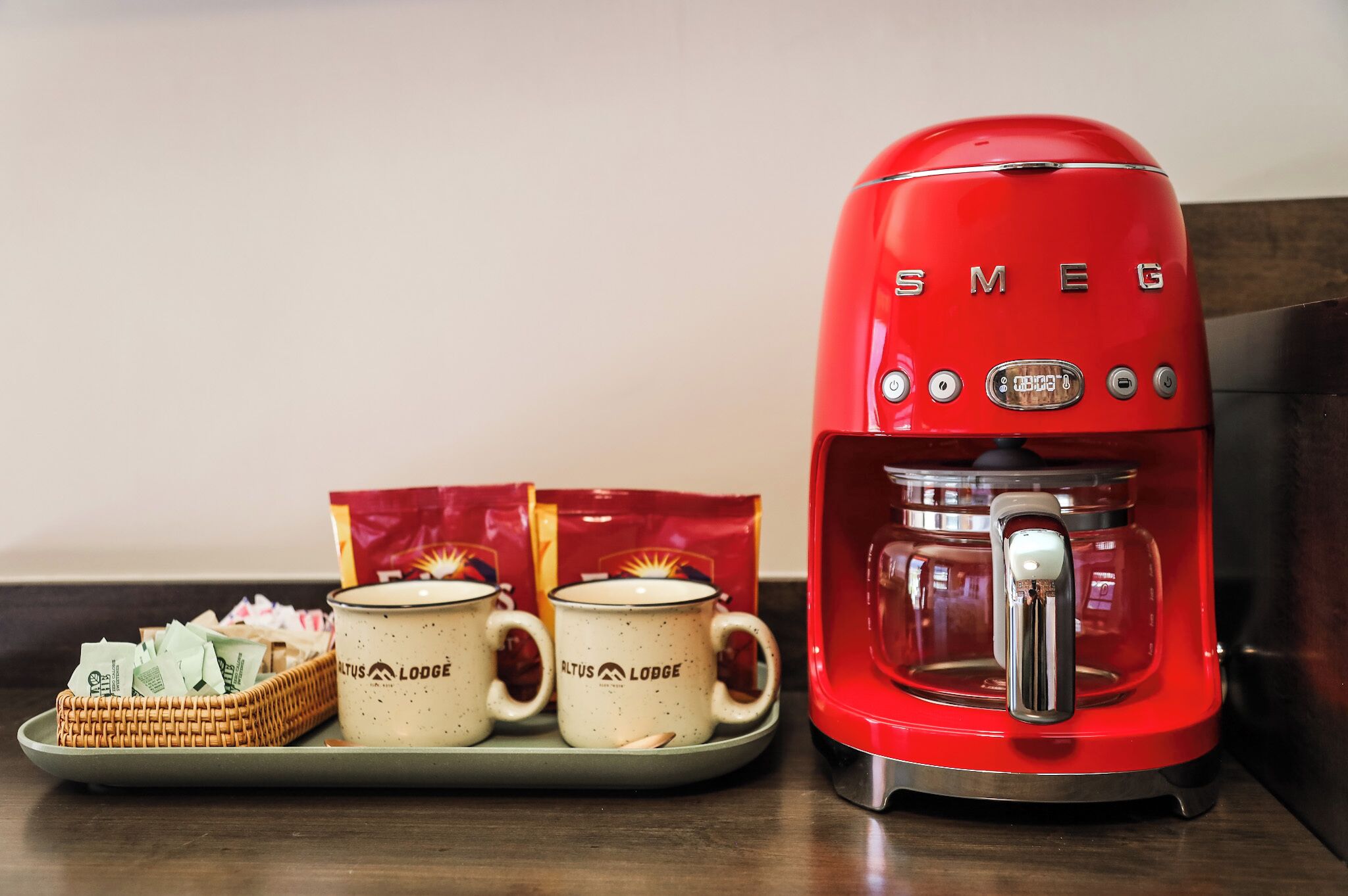 A red Smeg coffee maker with two mugs labeled "Altus Lodge" and a tray with a wicker basket and coffee packets.