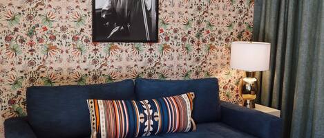 A living room with a blue sofa, patterned cushions, a framed picture, a lamp, and a small table.