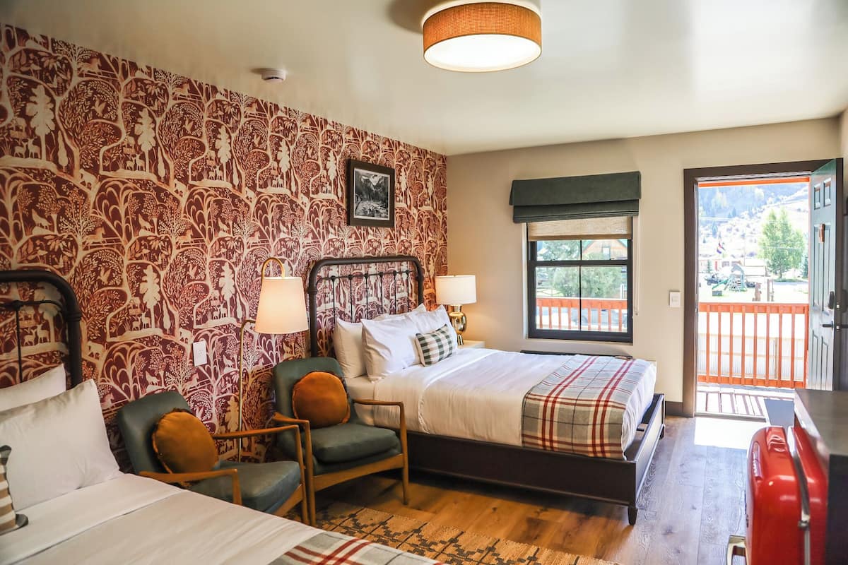 A bedroom with a patterned wallpaper, a bed with a plaid bedspread, two bedside lamps, a chair with a cushion, and a view of the outdoors through a window.