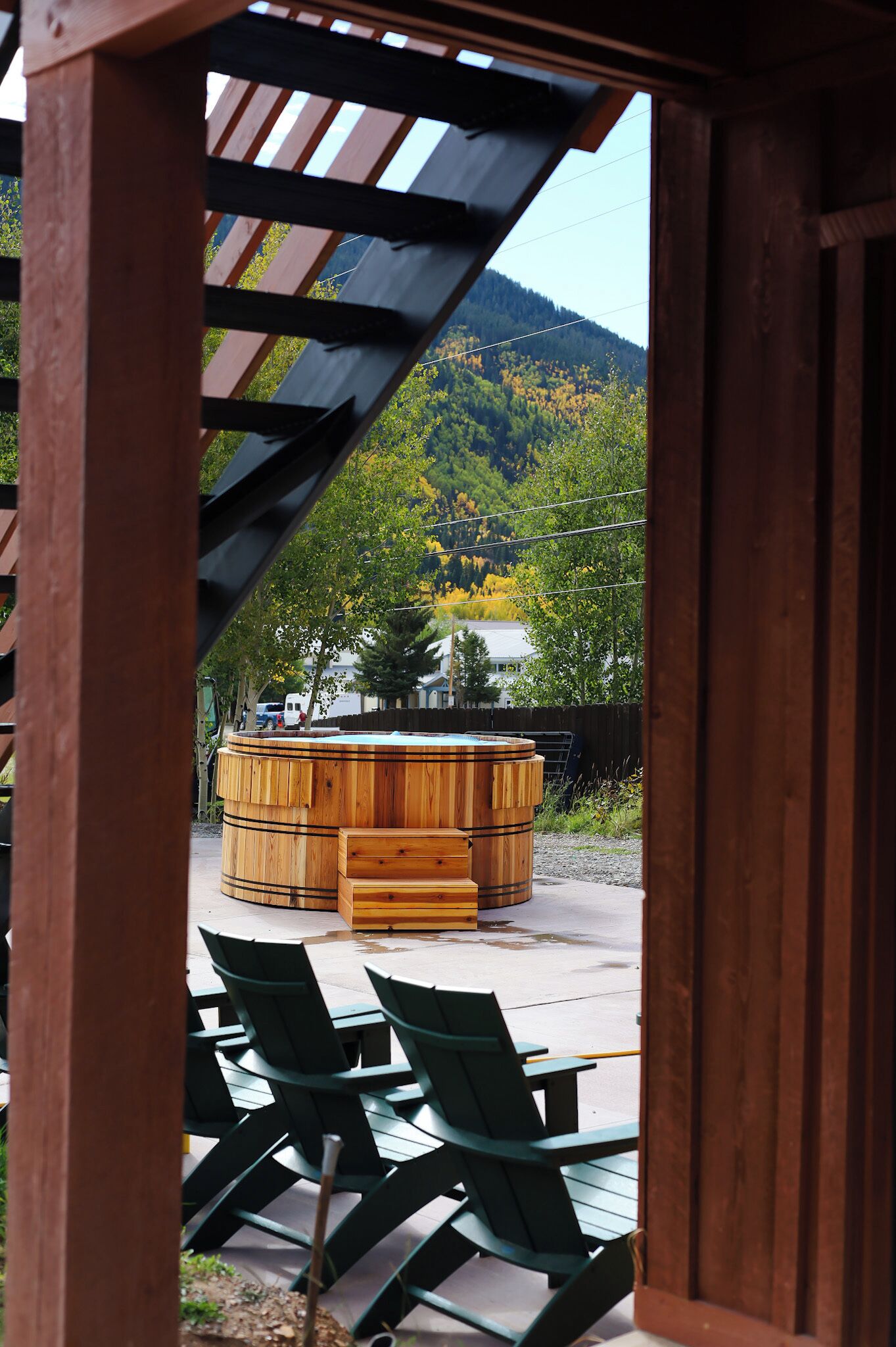 A wooden outdoor seating area with a wooden hot tub and chairs, set against a backdrop of a forested mountain.