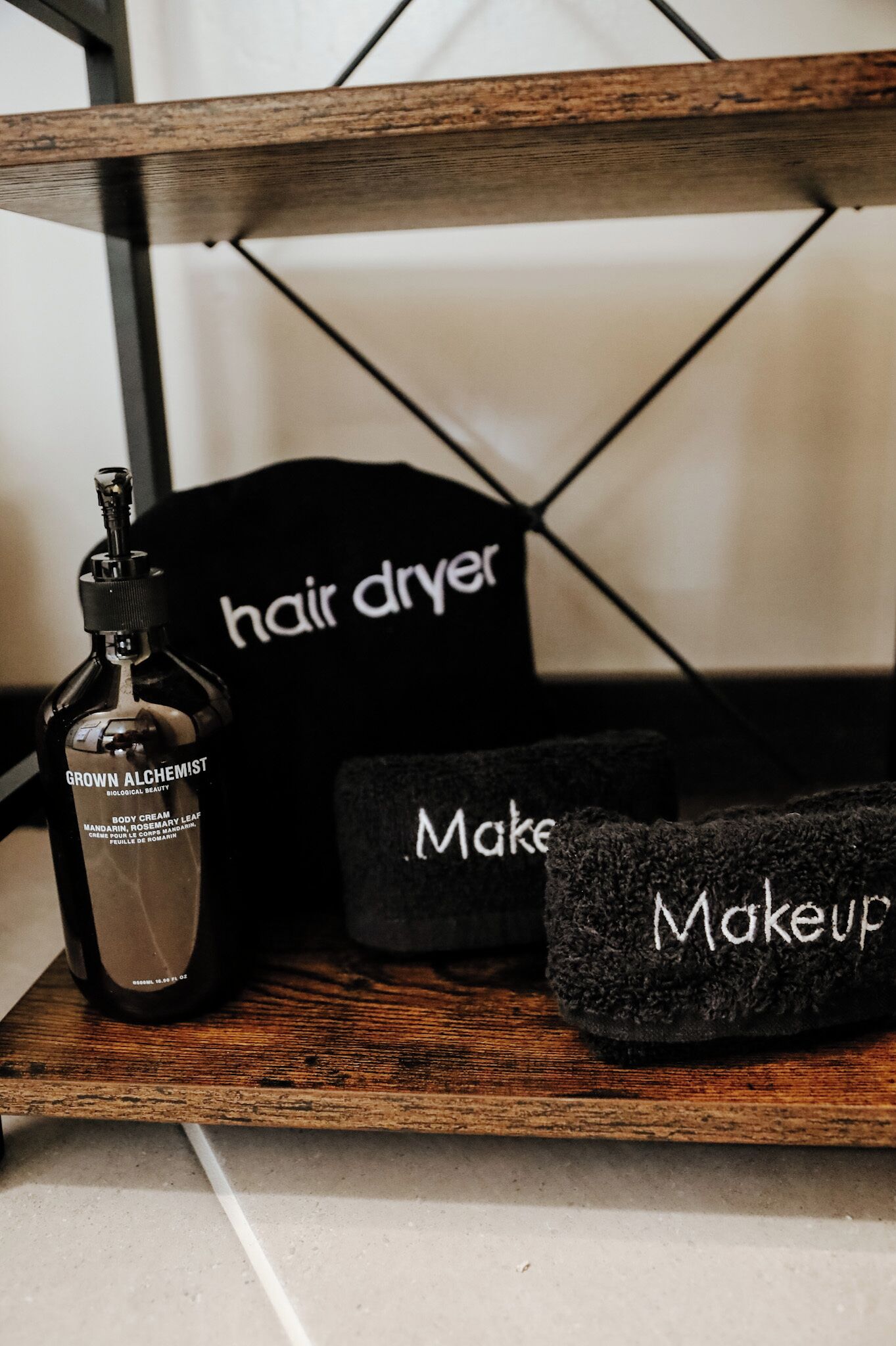 A wooden shelf with a bottle of 'Grown Alchemist' body cream, a black hair dryer, and two black makeup bags.