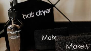 A wooden shelf with a bottle of 'Grown Alchemist' body cream, a black hair dryer, and two black makeup bags.