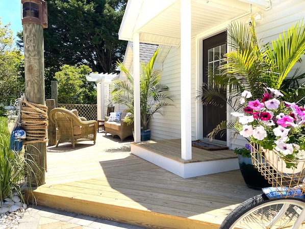 A wooden deck with a wicker chair and a small table, a potted plant with pink flowers, and a bicycle with a basket.