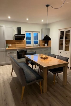 A modern kitchen with a dining area, featuring a wooden table, grey upholstered chairs, and a black pendant light.