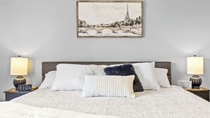 Una cama bien hecha con una manta blanca y estampada, dos lámparas de noche, un reloj digital y un cuadro con una imagen de un puente sobre un río con una torre.