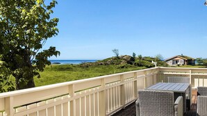 Eine Holzterrasse mit Korbmöbeln, einem weißen Geländer und Blick auf das Meer und die Hügel.