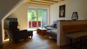A modern loft bedroom with a sloped ceiling, wooden beams, a large window, a sofa, a chair, and a bedside table. - Holiday Apartment Kieschtock (Unterkirnach)