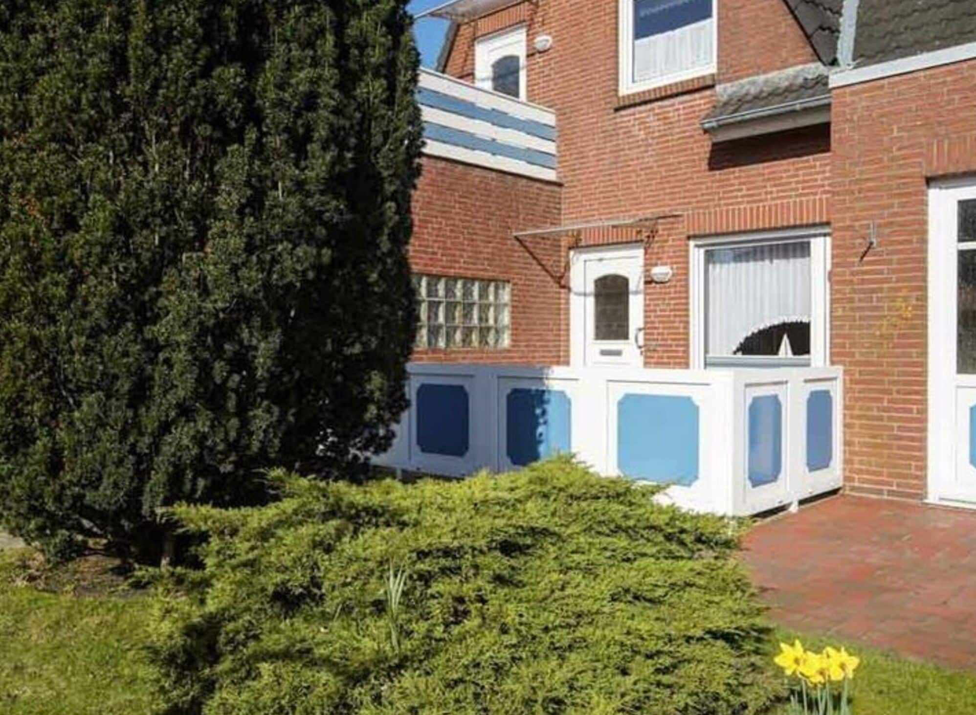 A two-story brick house with a white door and windows, surrounded by greenery and a small garden.