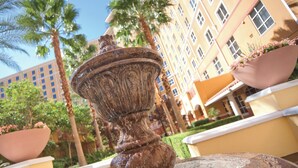 A fountain with water spouting from a central column, surrounded by potted plants and palm trees, with a building in the background. - Wyndham Grand Desert 2 BR Deluxe - Full Resort Access -25% Deposit to Book NOW! (Las Vegas)