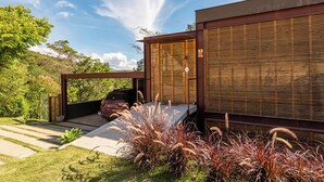 A modern house with a covered driveway, a parked car, and a lush garden. - Santa Serra Itaipava (Petrópolis)