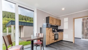 A modern kitchen with wooden cabinets and a built-in oven, a dining area with a table and chairs, and large windows offering a view of a forested area.