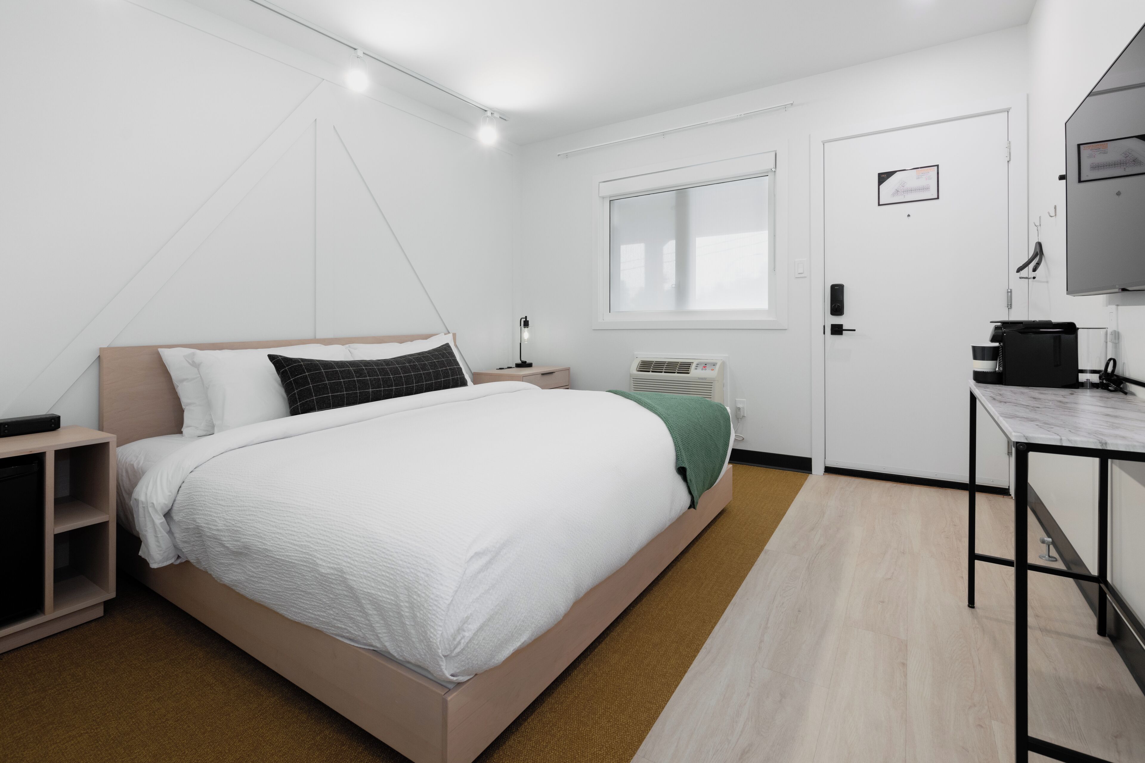 Habitació d'hotel moderna amb llit, taula de nit, làmpada, aparell d'aire condicionat i un petit escriptori amb cadira.