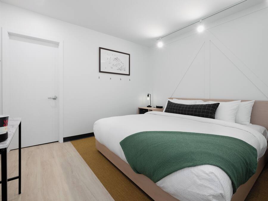A modern hotel room with a large bed, a bedside table, a nightstand, a desk with a chair, and a door.