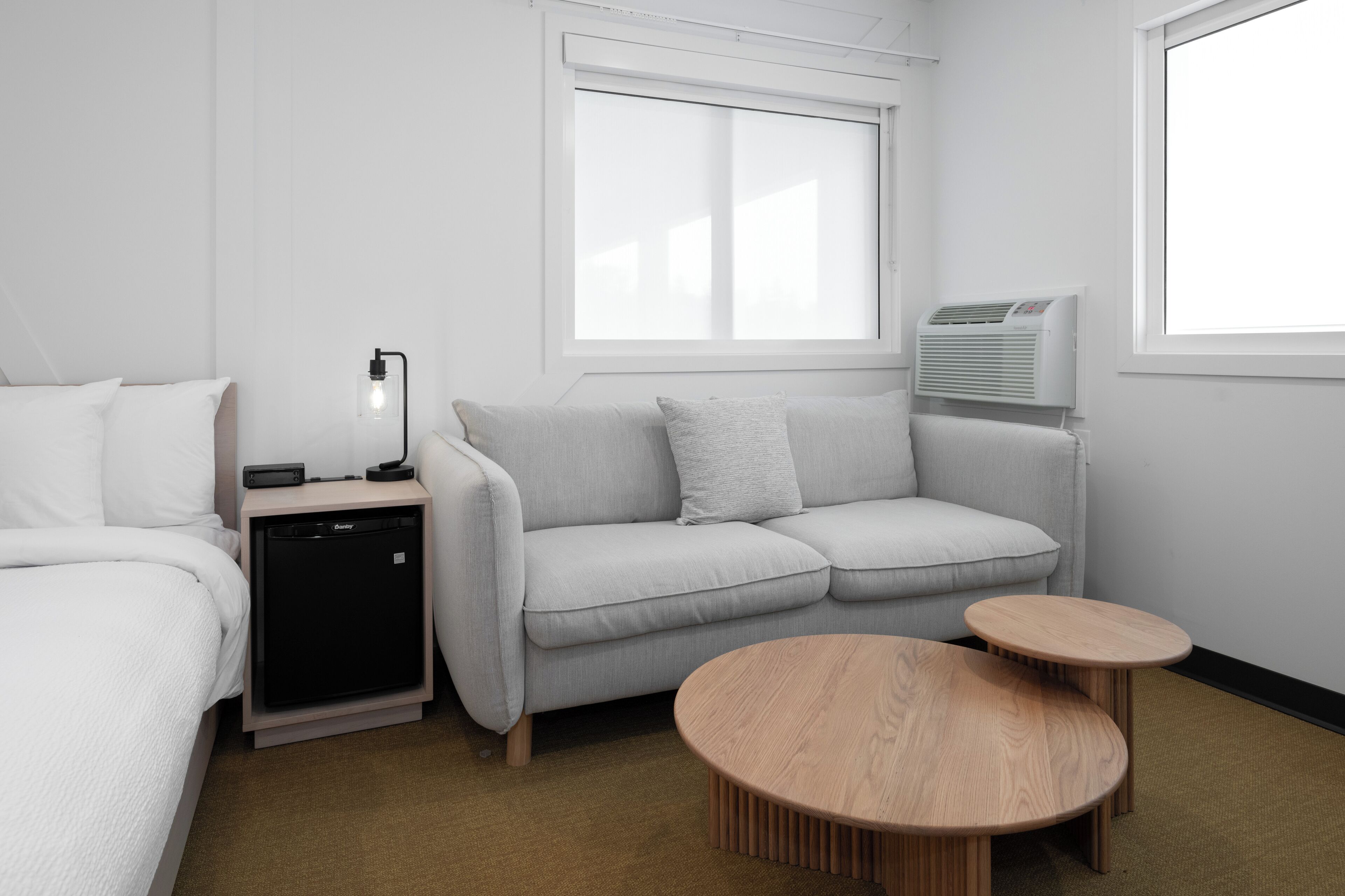 Ein modernes Hotelzimmer mit grauem Sofa, einem kleinen Holztisch, einem Nachttisch mit Lampe und einer kompakten Klimaanlage.