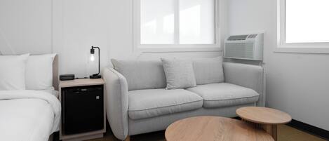 A modern hotel room with a grey sofa, a small wooden table, a bedside table with a lamp, and a compact air conditioning unit.