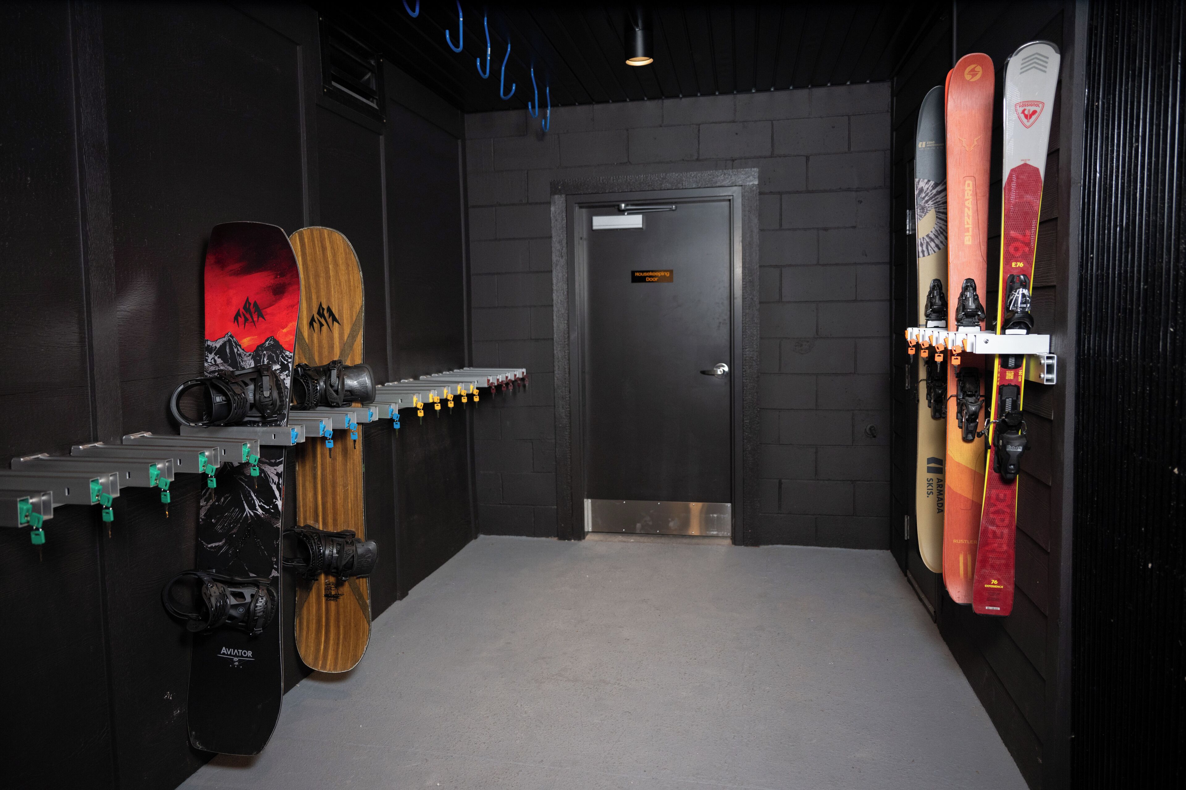 Een skiopslagruimte met ski's en snowboards op rekken, een deur op de achtergrond en een plafondlamp.