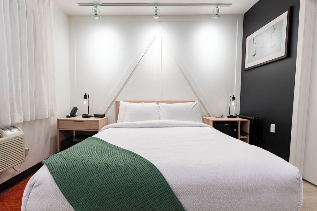 Una camera d'albergo con un letto, comodini, una lampada, un'opera d'arte appesa al muro e una finestra con tende.