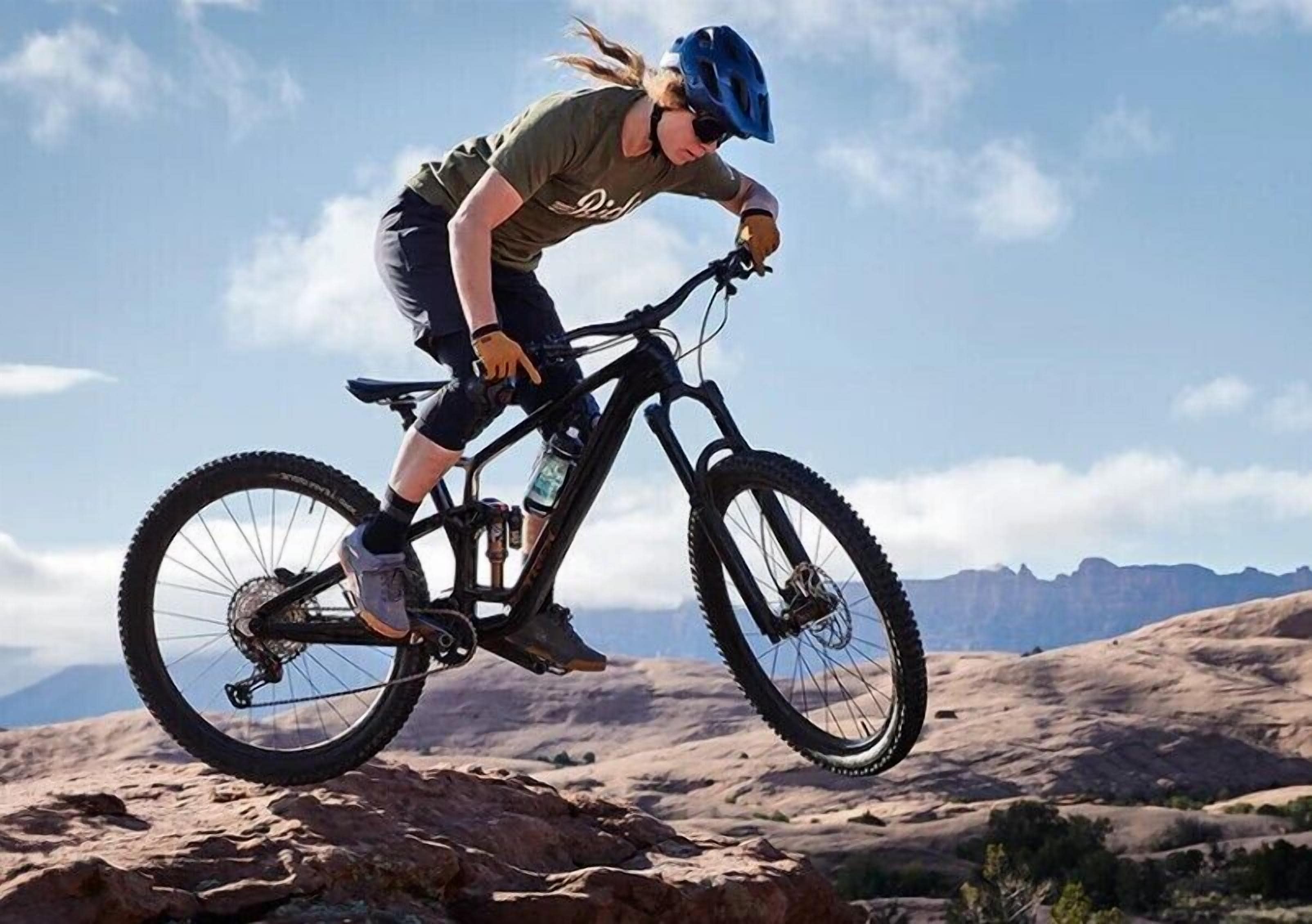 Una persona montando una bicicleta de montaña sobre terreno rocoso, con cielo despejado y montañas distantes al fondo.