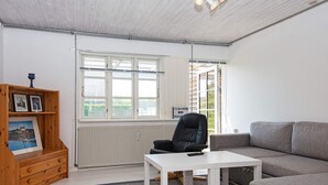 Una sala moderna con un sofá gris, una mesa blanca y una silla negra. Hay un mueble de madera con fotografías y un radiador en la pared.