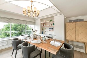 Uma sala de jantar com mesa de madeira posta para seis pessoas, rodeada por cadeiras, um lustre e vista para o verde lá fora.