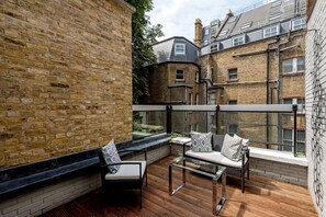 Un balcone con ringhiera in vetro, pavimento in legno e angolo relax con cuscini.