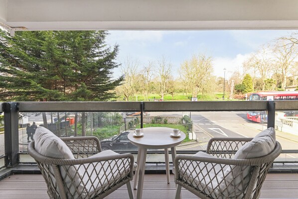 A balcony with wicker chairs and a small table, overlooking a parking area and trees. - Elegant Kensington Apartment · Hyde Park · Sleeps 4 (London)