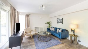 Una sala moderna con un sofá azul, una mesa de centro de vidrio, un sillón color beige y un televisor sobre un mueble negro.