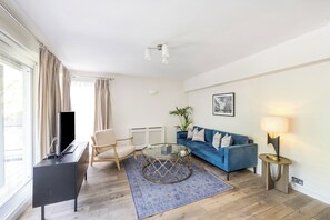 A modern living room with a blue sofa, a glass coffee table, a beige armchair, a TV on a black cabinet, and a lamp on a side table. - Elegant Kensington Apartment · Hyde Park · Sleeps 4 (London)