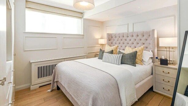 A bedroom with a large bed, a bedside table, a nightstand, a lamp, a radiator, and a window. - Stylish Kensington Apartment with Hyde Park View Terrace · Sleeps 4 (London)