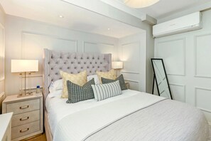 Un dormitorio ordenado con cabecera capitonada, una mesita de noche con lámpara, un espejo y un armario.