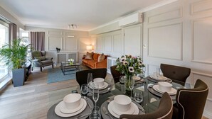 Un coin repas avec une table en verre dressée pour quatre personnes, entourée de chaises à l’assise rembourrée de couleur foncée. On aperçoit un canapé et une table basse avec une lampe à l’arrière-plan.