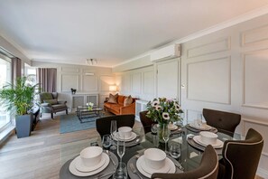 Uma sala de jantar com mesa de vidro posta para quatro pessoas, rodeada por cadeiras estofadas escuras. Um sofá e uma mesa de centro com abajur são visíveis ao fundo.