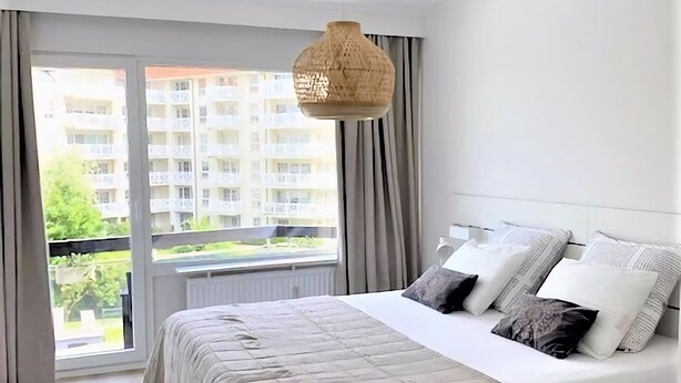 Une chambre avec un grand lit, un luminaire suspendu et une vue sur un immeuble d’habitation par la fenêtre.