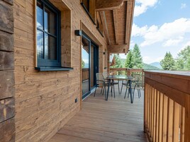 Terraza de madera con una mesa y sillas de vidrio, con vista a un área boscosa.