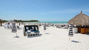 En strand med liggestole og parasoller, en stråtækt hytte og et poolområde.