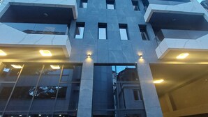 A modern building with a geometric facade, multiple balconies, and a parking area with cars and potted plants.
