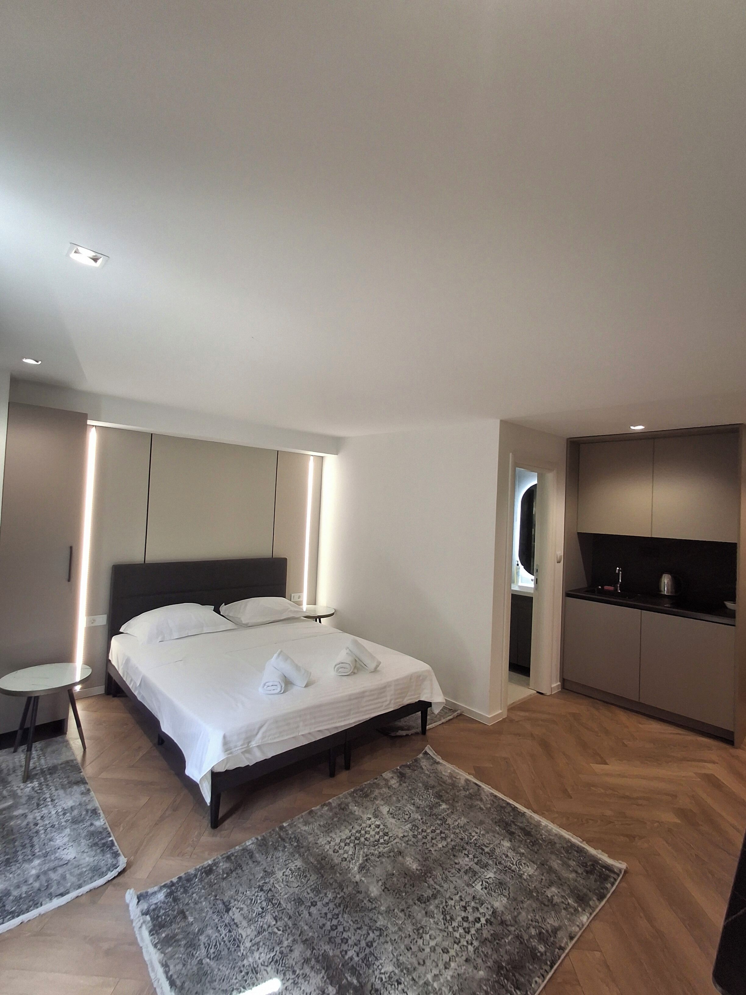 A modern hotel room with a bed, bedside tables, a small round table, a kitchen area, and a TV.
