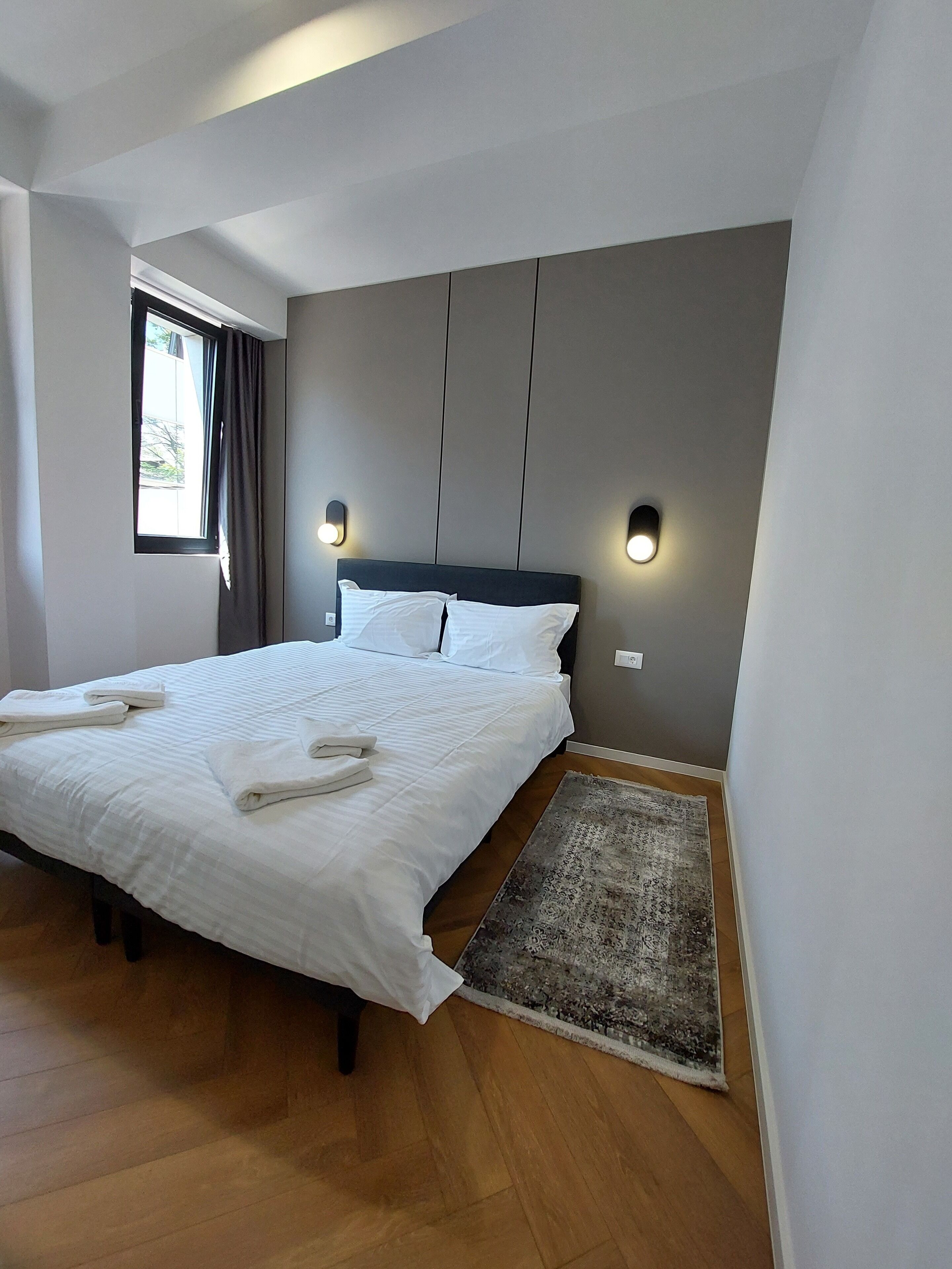 A modern bedroom with a large bed, two wall-mounted lights, a window with curtains, and a wooden floor.