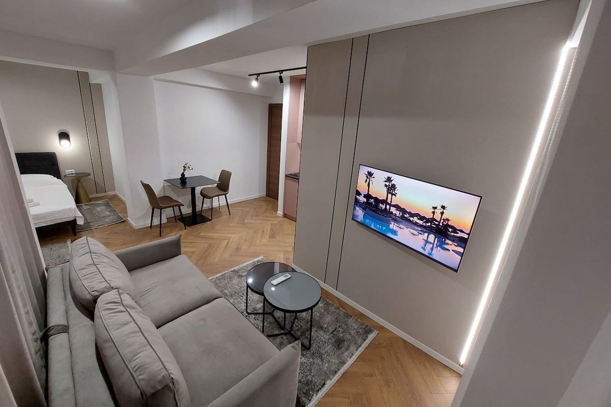 A modern hotel room with a sofa, a dining area, a bed, and a TV displaying a sunset scene.