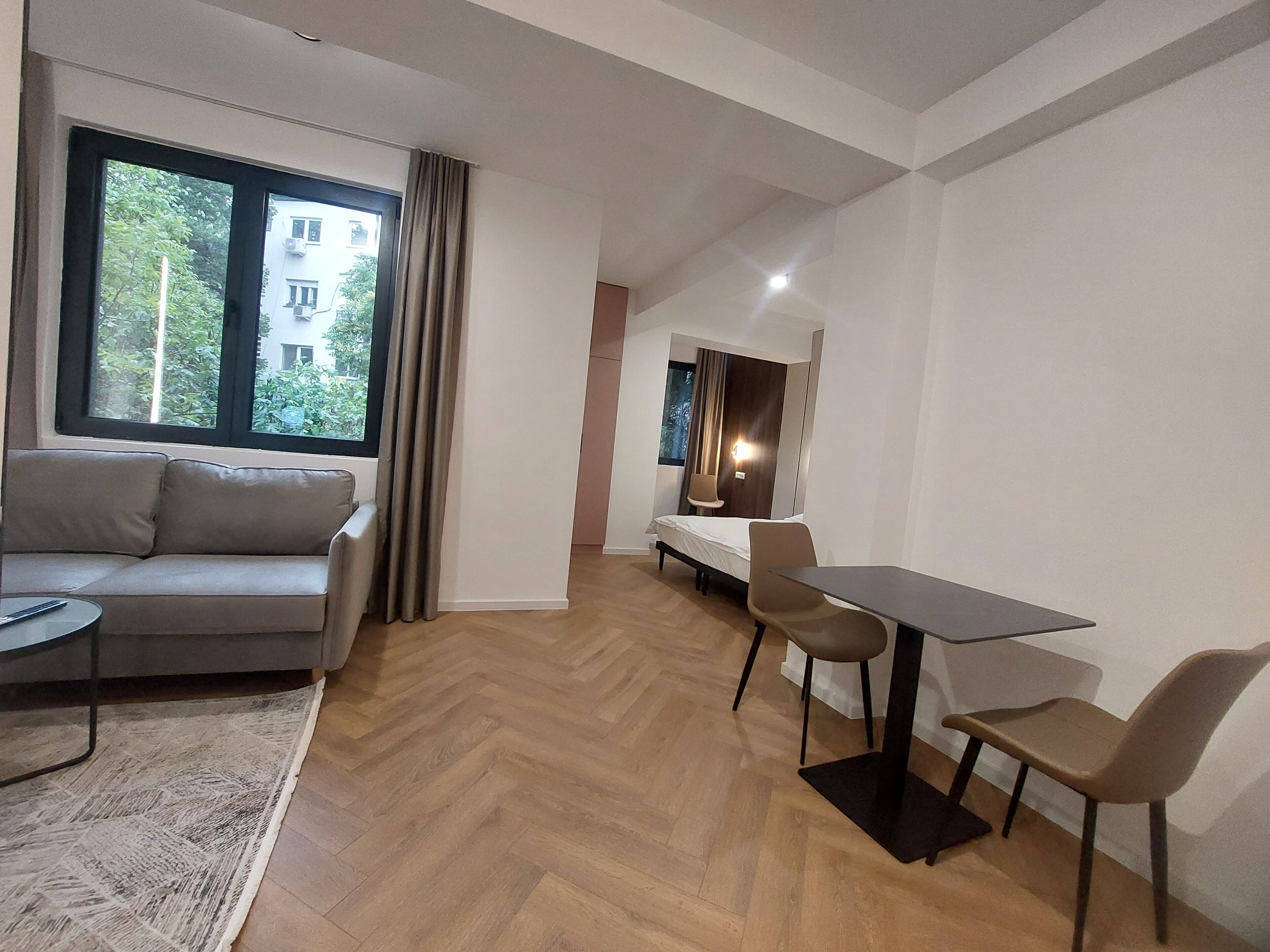 Une pièce moderne comprenant un canapé, une table à manger et des chaises. On y trouve une fenêtre avec des rideaux, une lampe et une vue sur la verdure à l’extérieur.